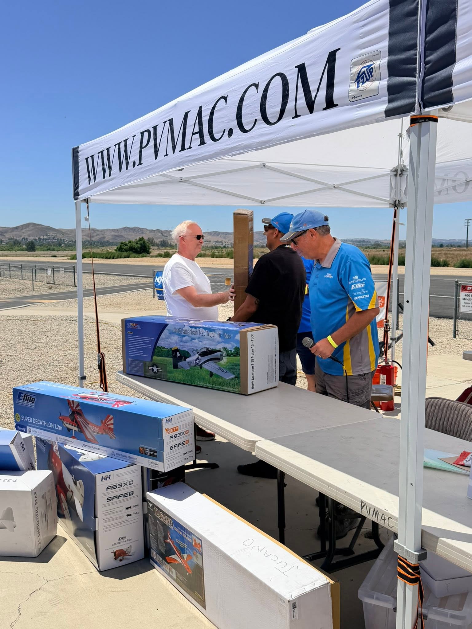 PVMAC members under the club tent with WWW.PVMAC.COM banner and RC airplane kit boxes on display